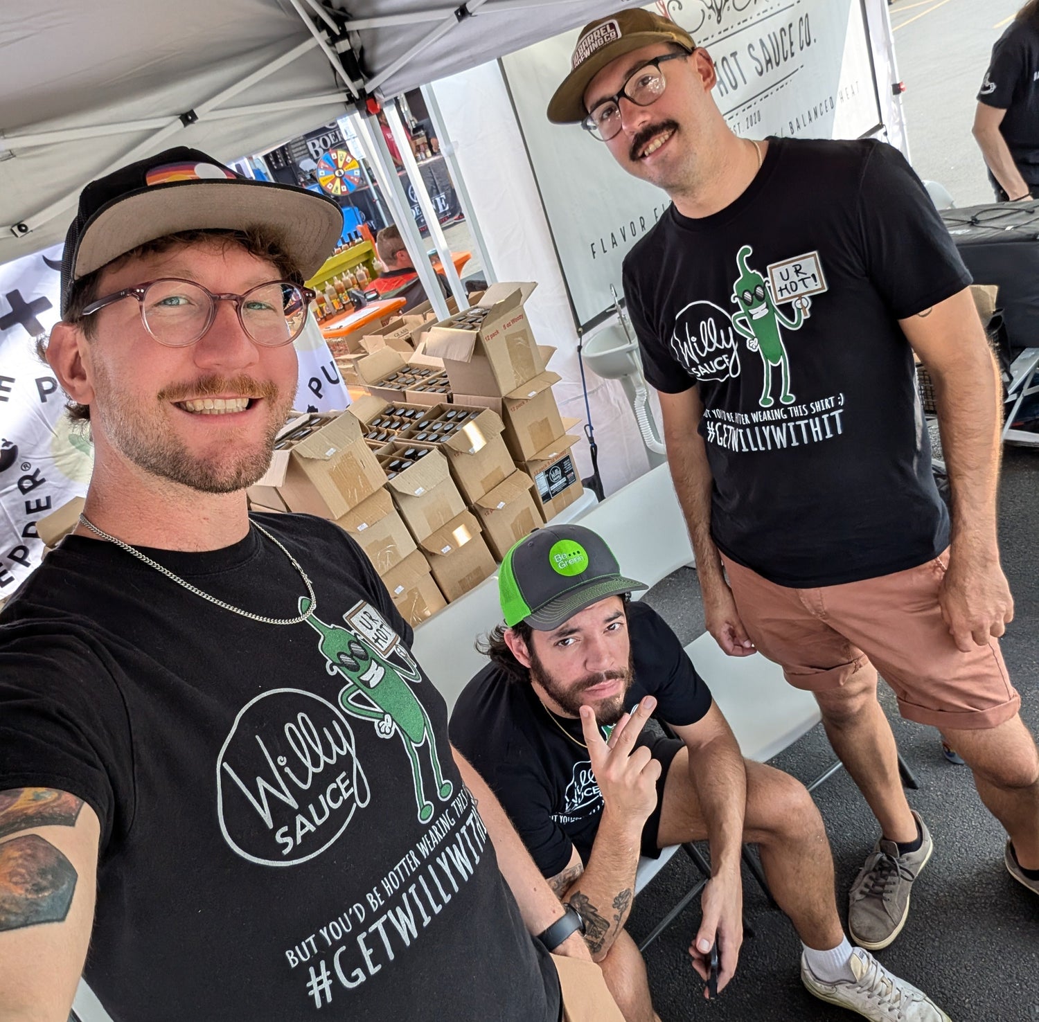 Three men wearing black 'Willy Sauce' t-shirts posing for a selfie under a tent.